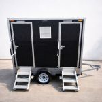Luxury Portable RestroomBlack portable restroom trailer with dual doors, aluminum steps, diamond plate trim and tow hitch parked outdoors beside a swimming pool